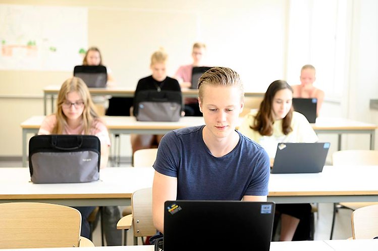 Elever sitter vid varsin dator och gör ett prov i ett glest möblerat klassrum. 